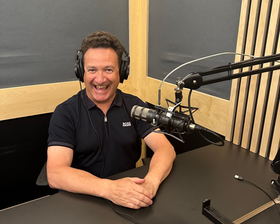 man smiling in a recording studio wearing headphones sitting at a desk with a microphone broadcasting a joyful podcast session